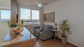 A modern living room with a grey sofa, a dining table with a fruit bowl, a ceiling fan, and a large window with a view. - Arena MRV, 2 quartos,vaga | Brazilian Corner (Belo Horizonte)