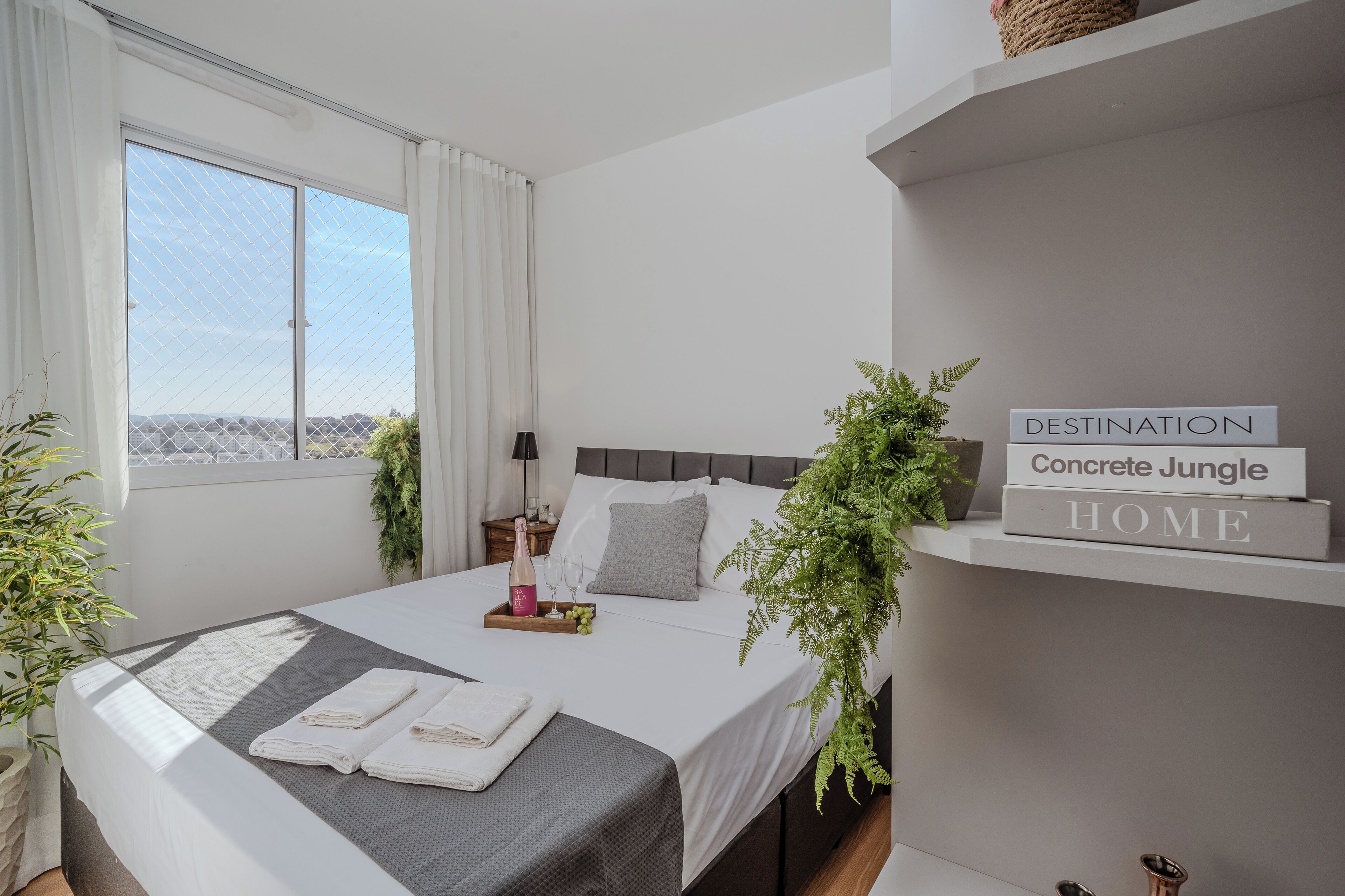 A modern bedroom with a bed, bedside table, and a window with curtains. There are two potted plants, a bedside lamp, and a tray with a bottle and glasses on the bed.