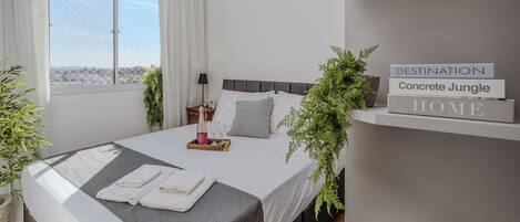A modern bedroom with a bed, bedside table, and a window with curtains. There are two potted plants, a bedside lamp, and a tray with a bottle and glasses on the bed.