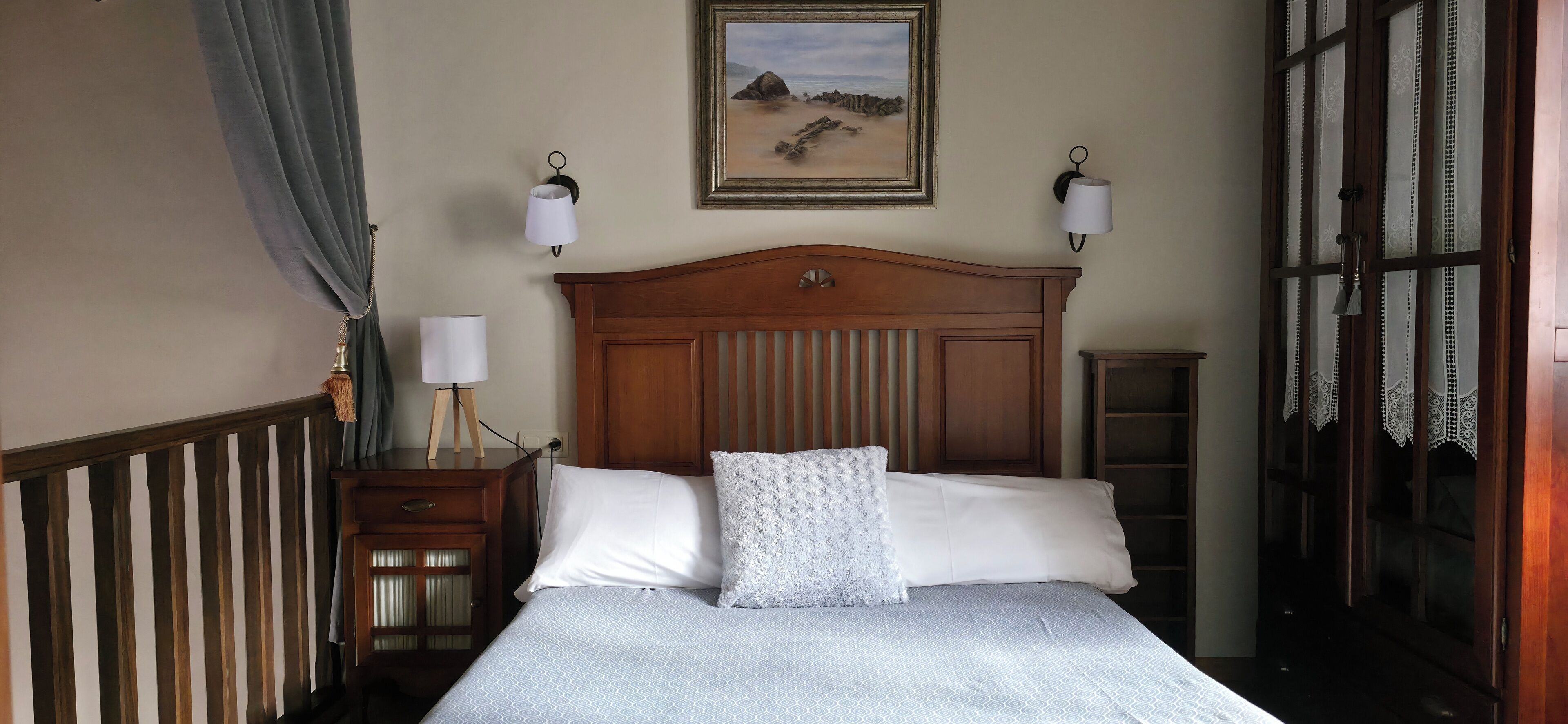 A neatly made bed with a patterned bedspread and two pillows, a wooden headboard, a nightstand with a lamp, and a framed picture on the wall.