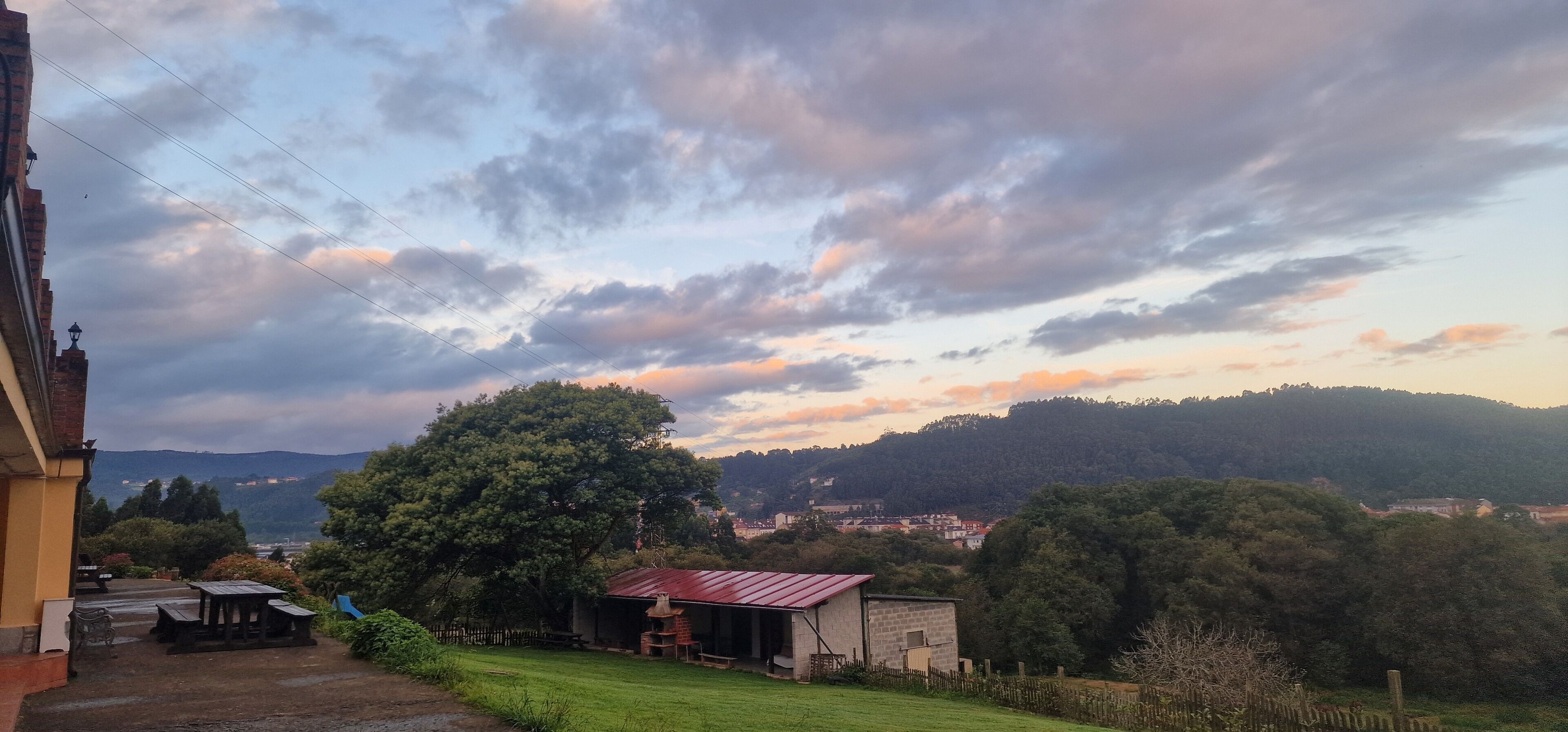 Paisaje rural con vista a un pueblo, un árbol grande y un edificio pequeño con techo rojo.