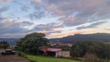 Paisaje rural con vista a un pueblo, un árbol grande y un edificio pequeño con techo rojo.