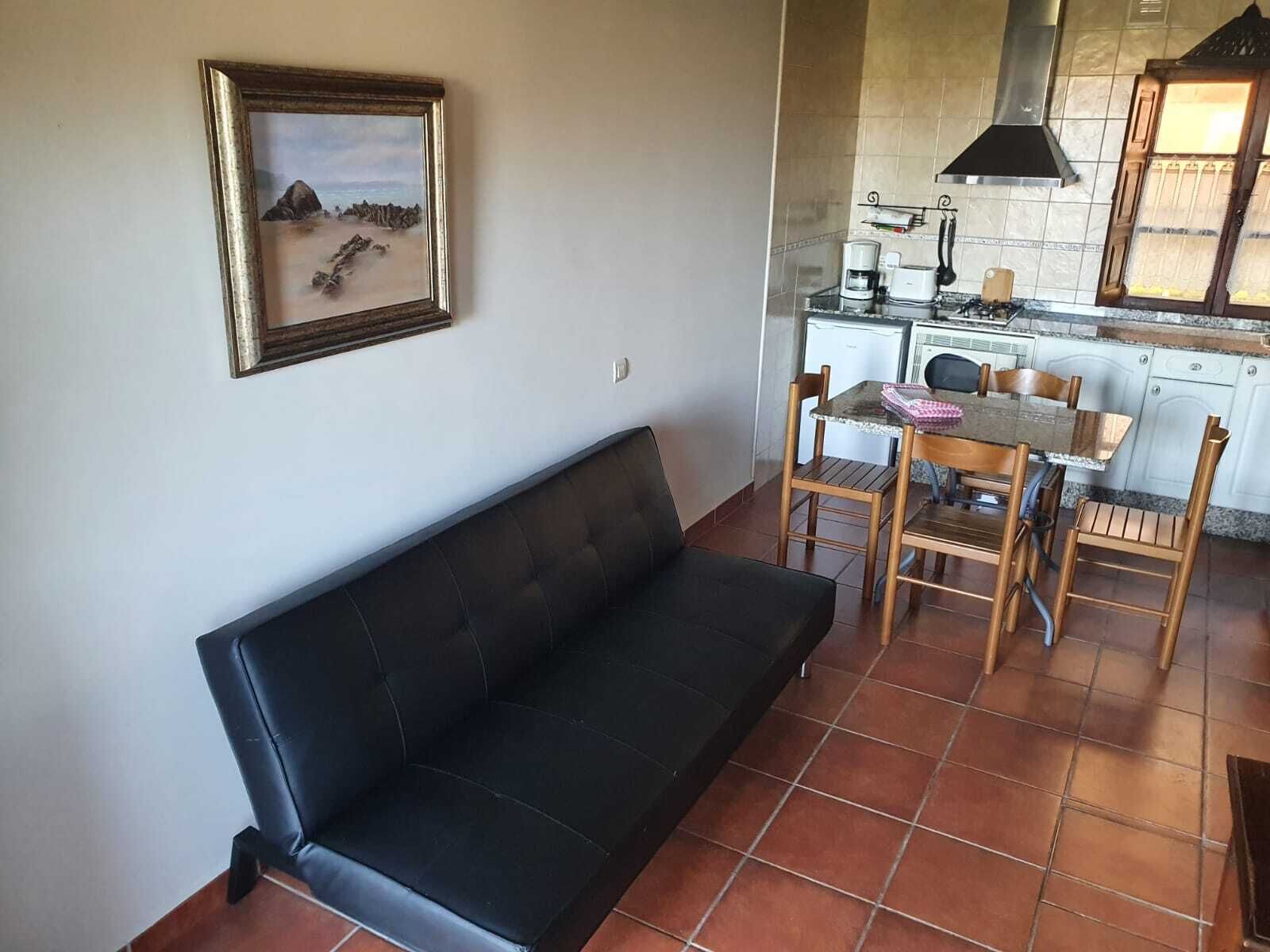 A living area with a black sofa, a dining table with chairs, a kitchen with a stove, and a framed picture on the wall.