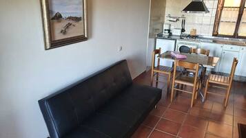 A living area with a black sofa, a dining table with chairs, a kitchen with a stove, and a framed picture on the wall.