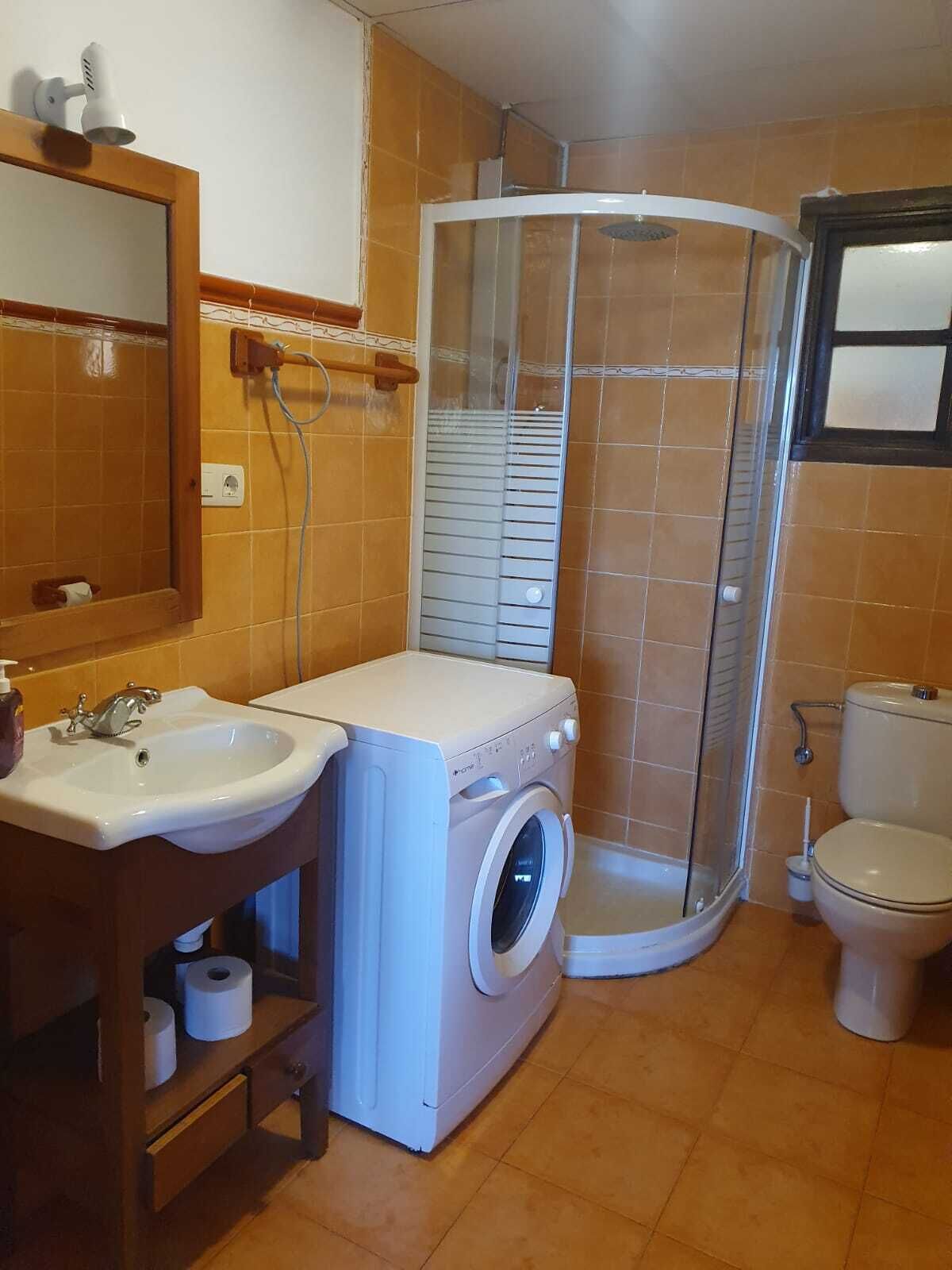 A compact bathroom with a sink, washing machine, toilet, and a glass shower enclosure.