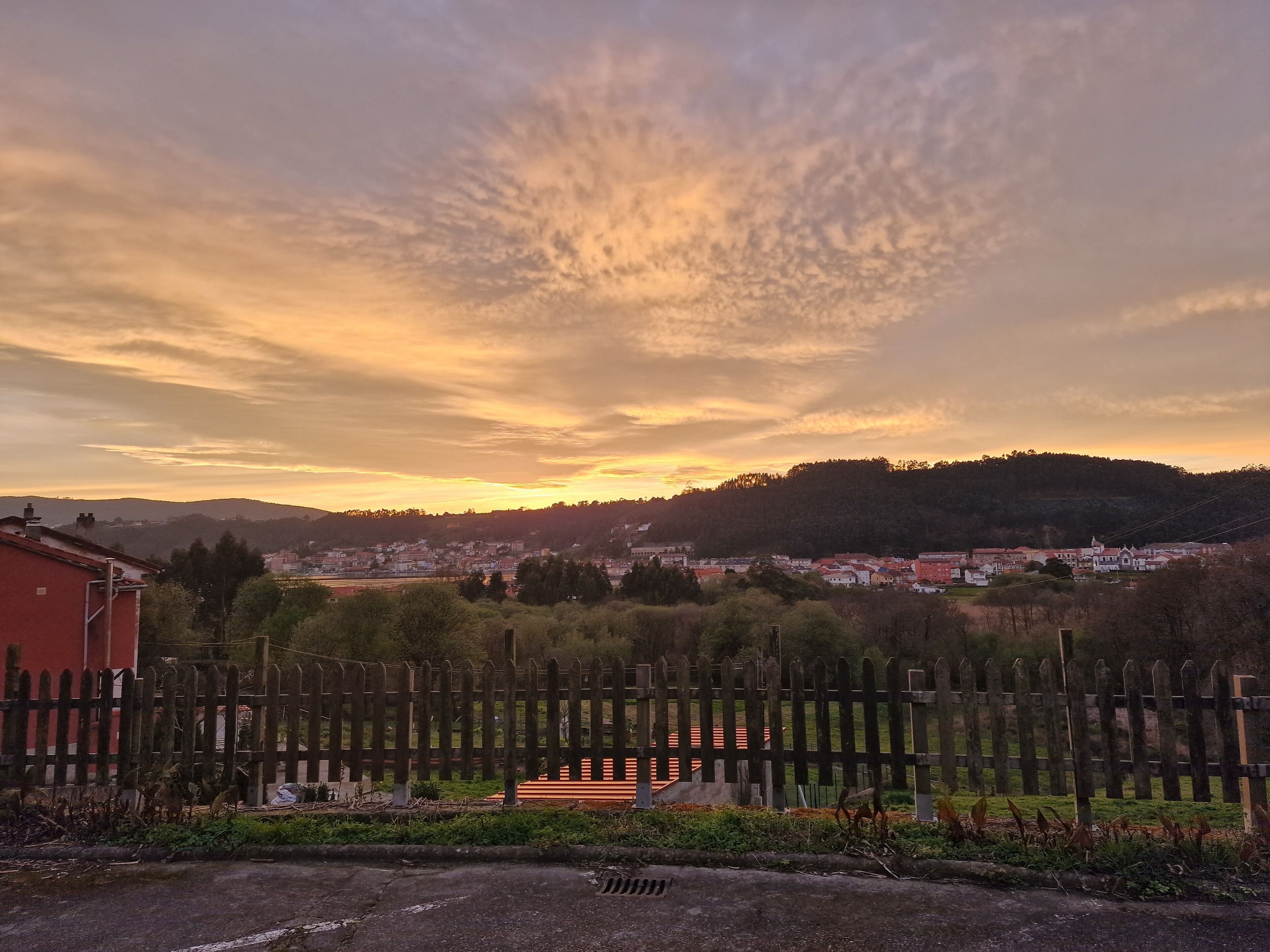 A rural landscape with a wooden fence, a small building, and a distant town under a sunset sky.