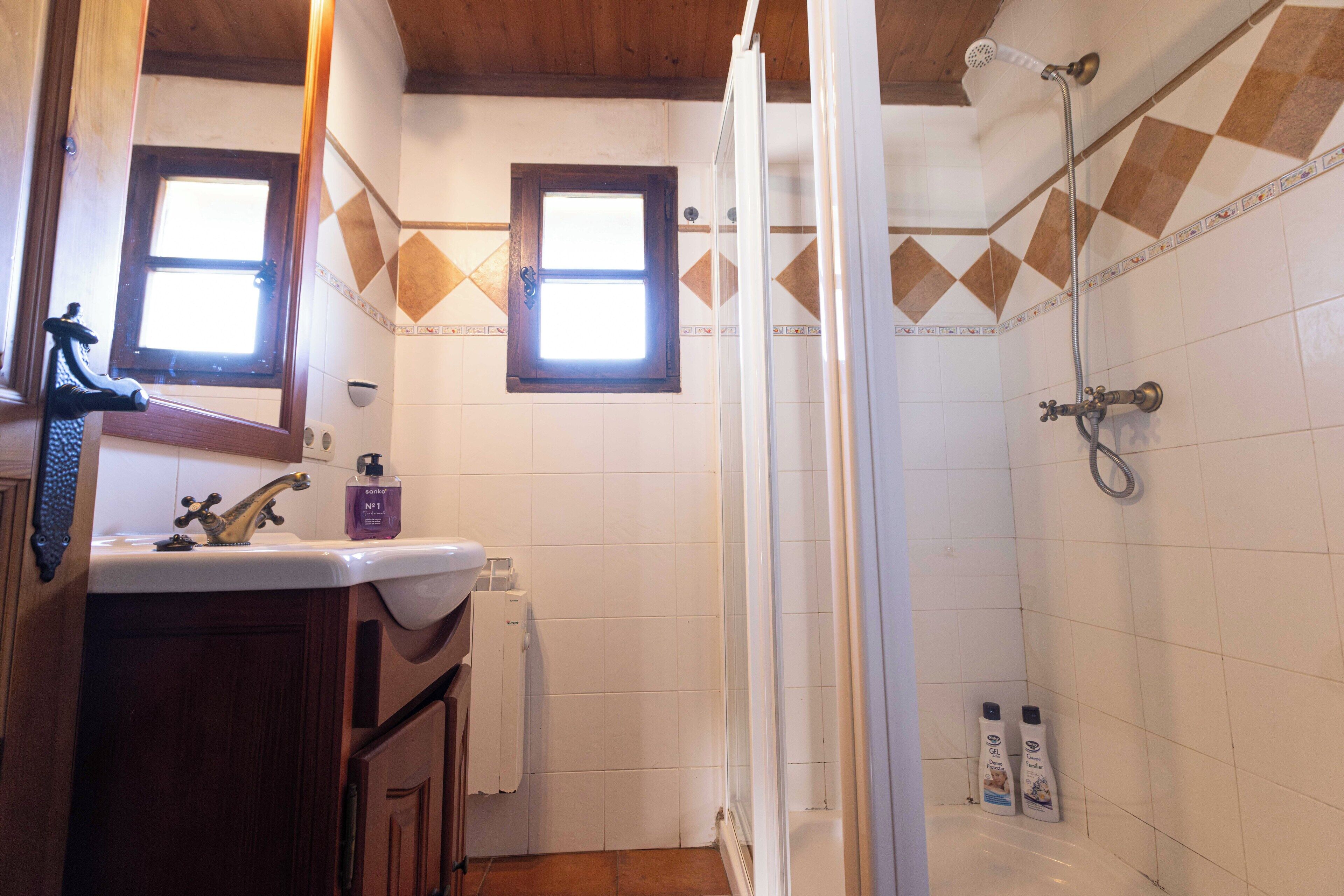 A bathroom with a sink, mirror, window, shower, and toiletries.