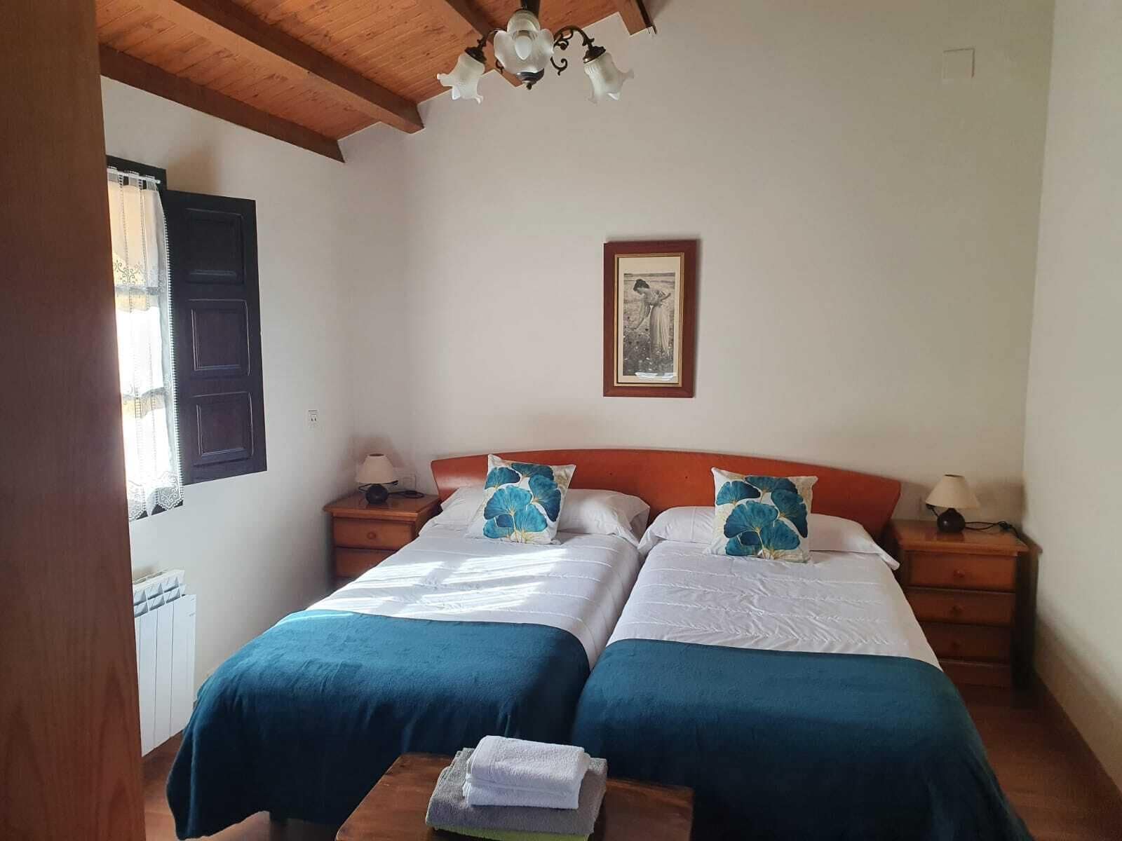 Una habitación doble con dos camas, cabeceros de madera, mesitas de noche con lámparas, un cuadro en la pared y una ventana con cortinas.