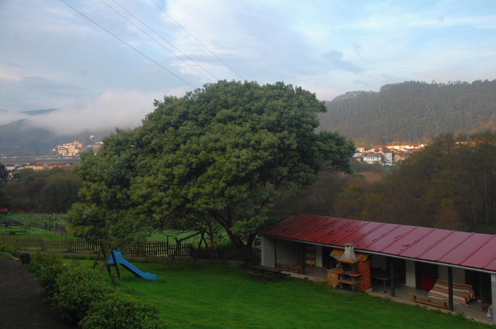 Un paisaje rural con un árbol grande, un edificio de techo rojo y un tobogán azul en un área con césped.