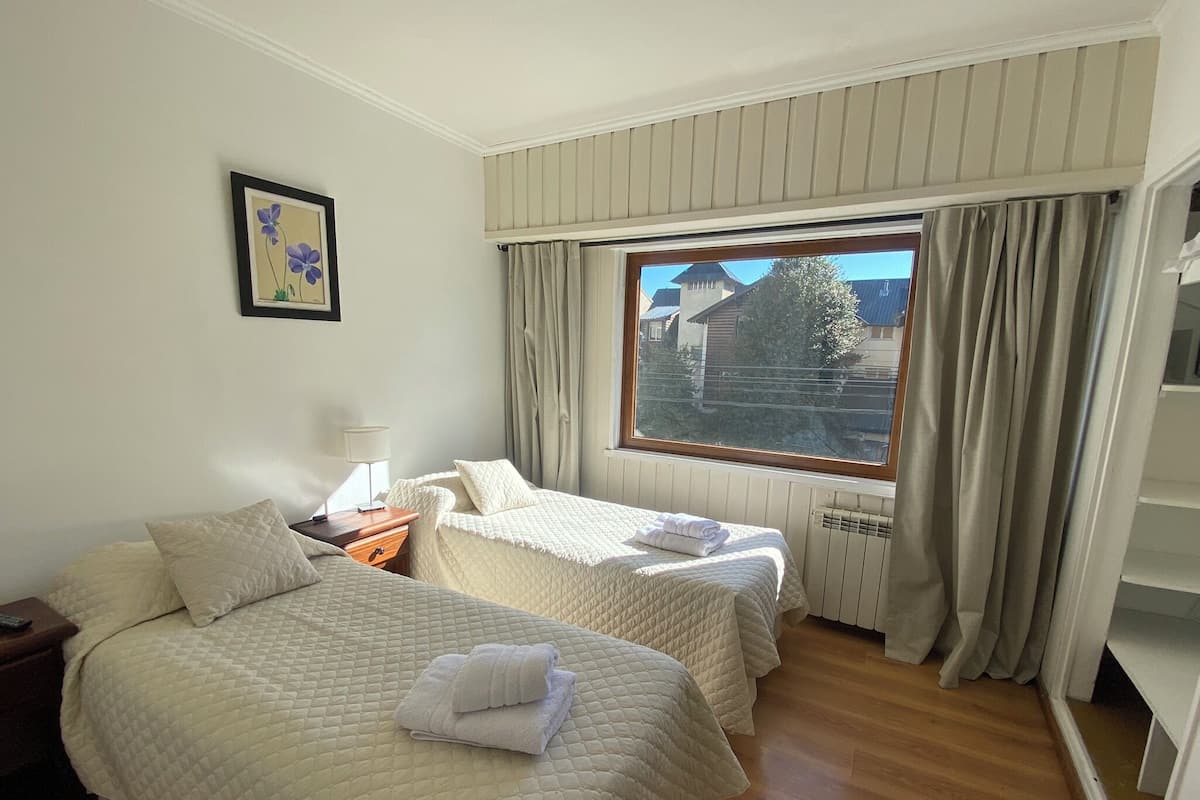 A hotel room with two beds, a window with curtains, a radiator, and a bedside table with a lamp.
