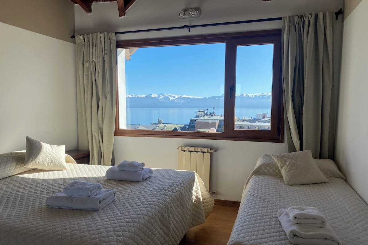 A hotel room with two beds, a view of mountains and a body of water through a large window, and a radiator on the wall.