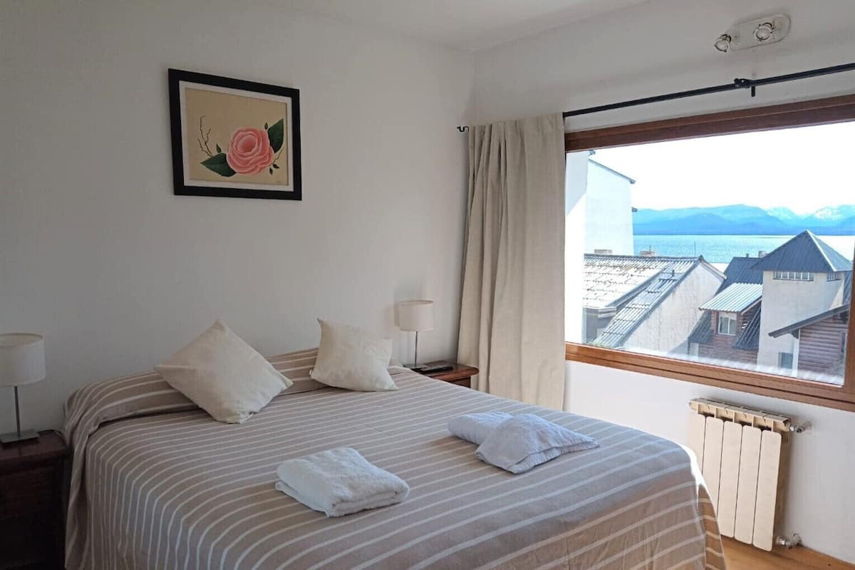 A bedroom with a bed, pillows, a bedside table with a lamp, a framed picture on the wall, and a window with a view of buildings and mountains.