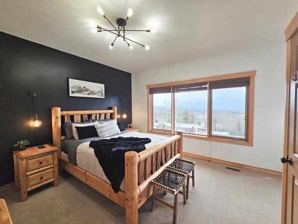 A bedroom with a wooden bed, a nightstand, a bench, a large window with a view, and a ceiling light fixture.
