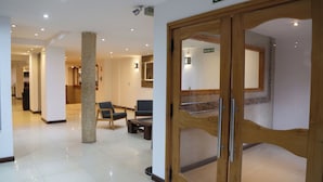 Un hall de hotel con una puerta de madera que indica 'SALIDA', suelo de baldosas y zonas de estar con sillas y mesas.