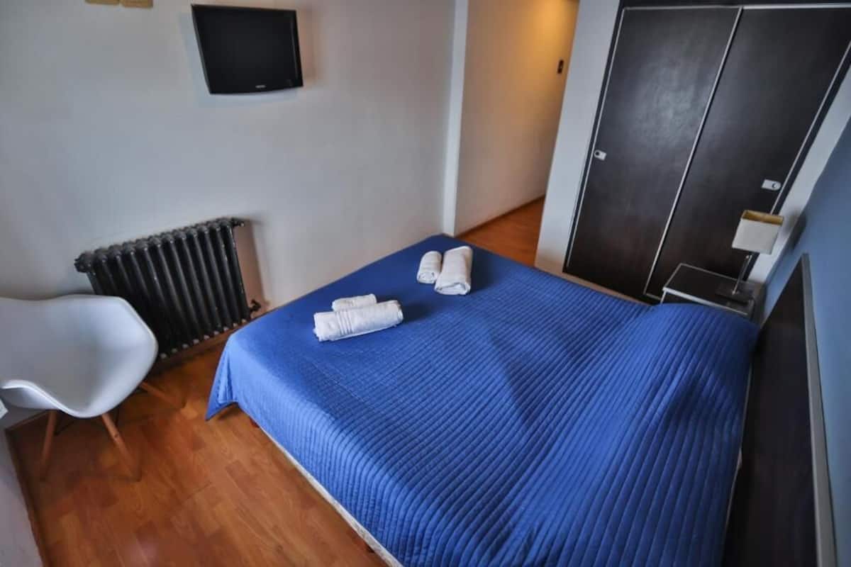 A small, neatly arranged room with a bed, a chair, a radiator, a television, and a wardrobe.