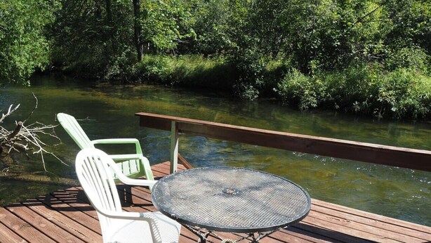 A wooden deck with a round table and two white chairs facing a river. - Sleeping Bear Riverside Cabins - Cabin #3 (Honor)