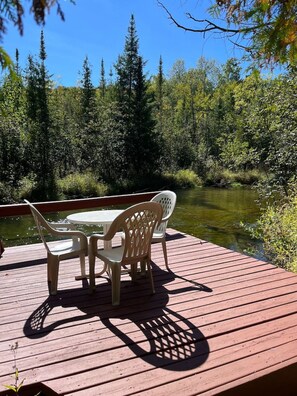 A wooden deck with a table and chairs overlooking a river, surrounded by dense forest. - Sleeping Bear Riverside Cabins - Cabin #2 (Honor)
