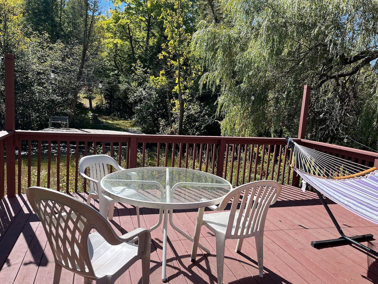 Una terraza de madera con una mesa y sillas de vidrio, una hamaca, y vistas a árboles y una masa de agua.
