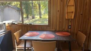 A wooden dining table with four chairs, a window with a view of trees and a house, and a wooden wall panel. - Sleeping Bear Riverside Cabins - Cabin #1 (Honor)