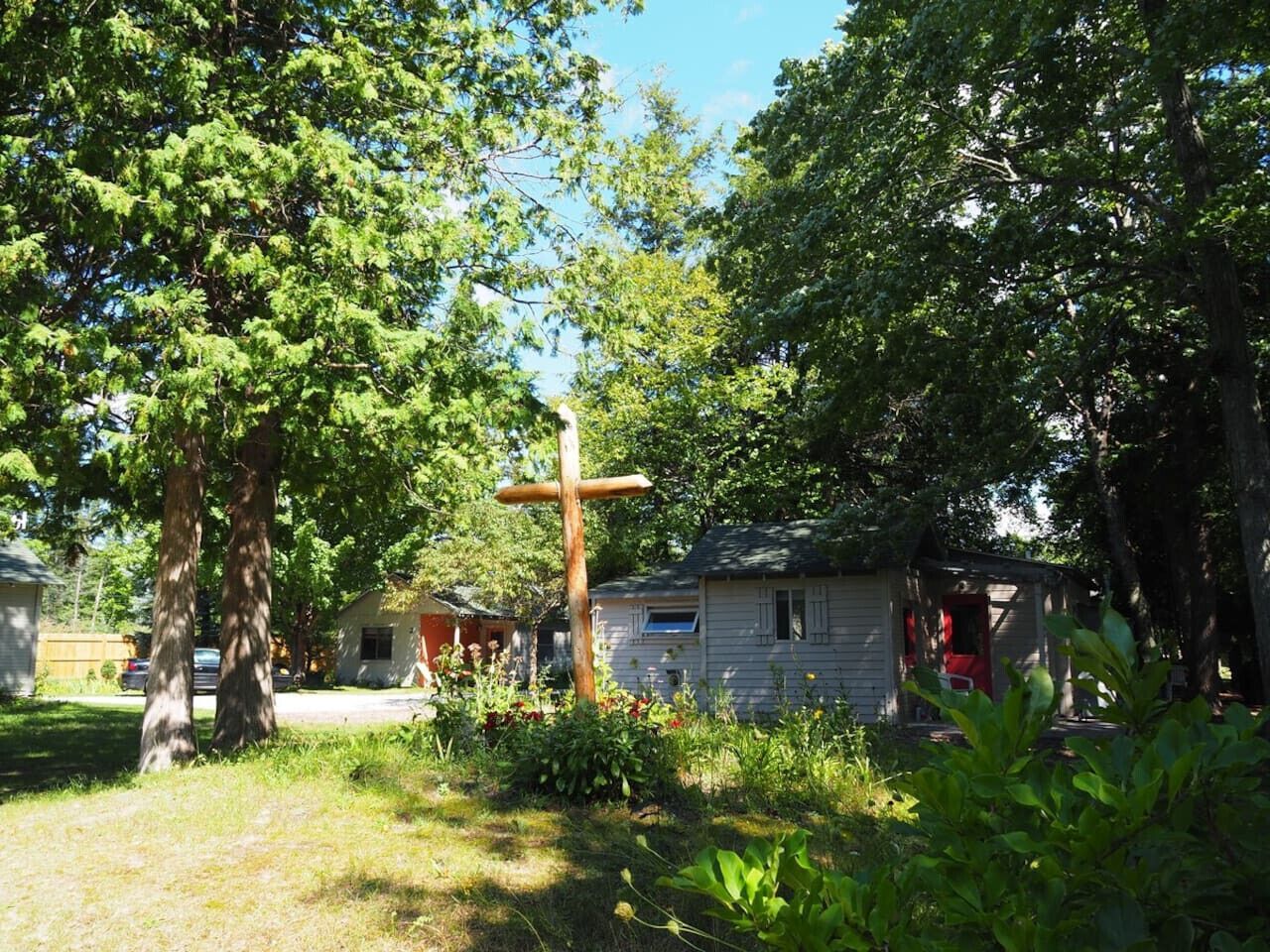 A rustic cabin with a wooden cross, surrounded by lush greenery and a clear blue sky.
