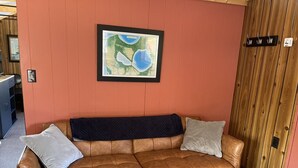 A brown leather sofa with two pillows against a red wall, a framed map, and a wooden door with hooks. - Sleeping Bear Riverside Cabins - Cabin #1 (Honor)