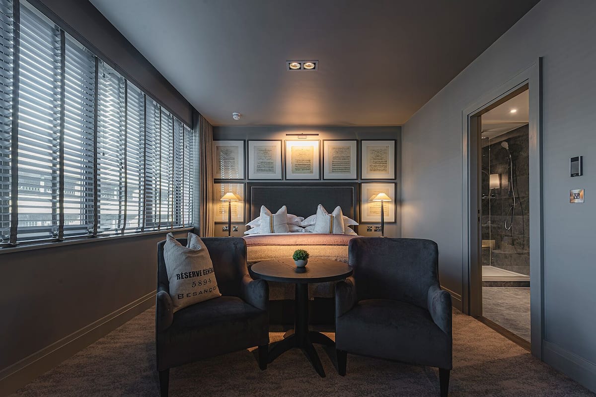 Una habitación de hotel con una cama grande, dos sillones, una mesita y una ventana con persianas.