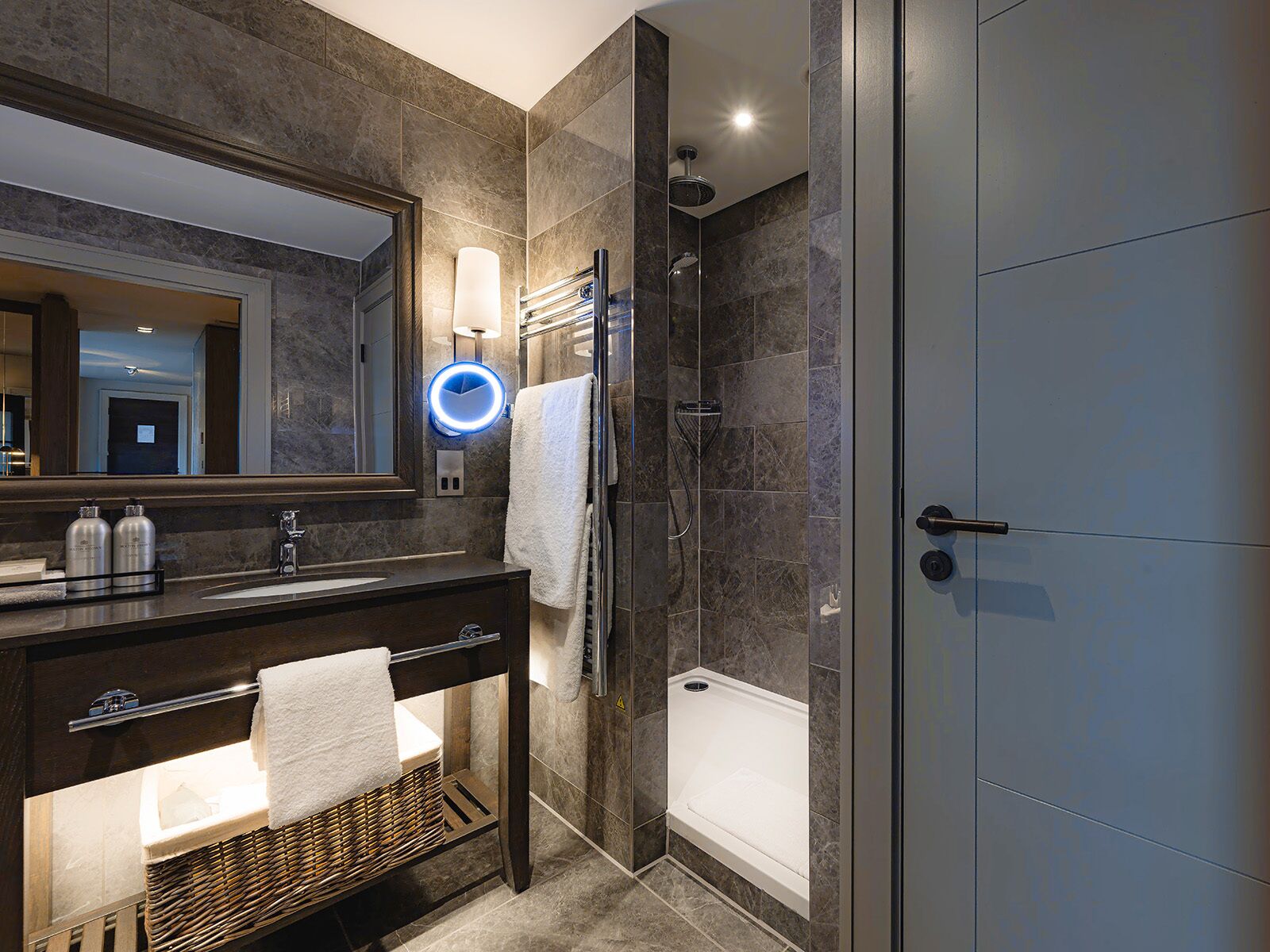 Un baño moderno con pared de mármol oscuro, un espejo grande, una lámpara redonda, una bañera blanca y una ducha con cerramiento de vidrio.