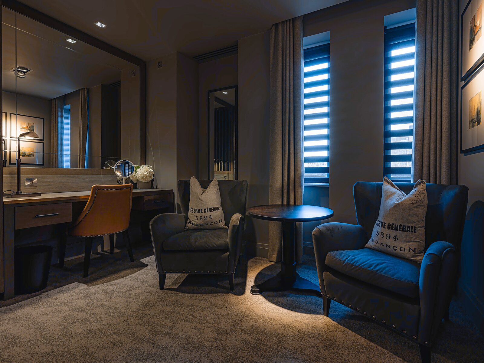 A hotel room with a desk, two armchairs, a round table, a mirror, and a large window with blinds.