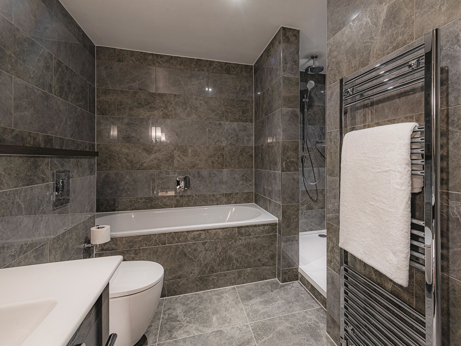 A modern bathroom with a bathtub, toilet, and shower area, featuring dark marble tiles and chrome fixtures.