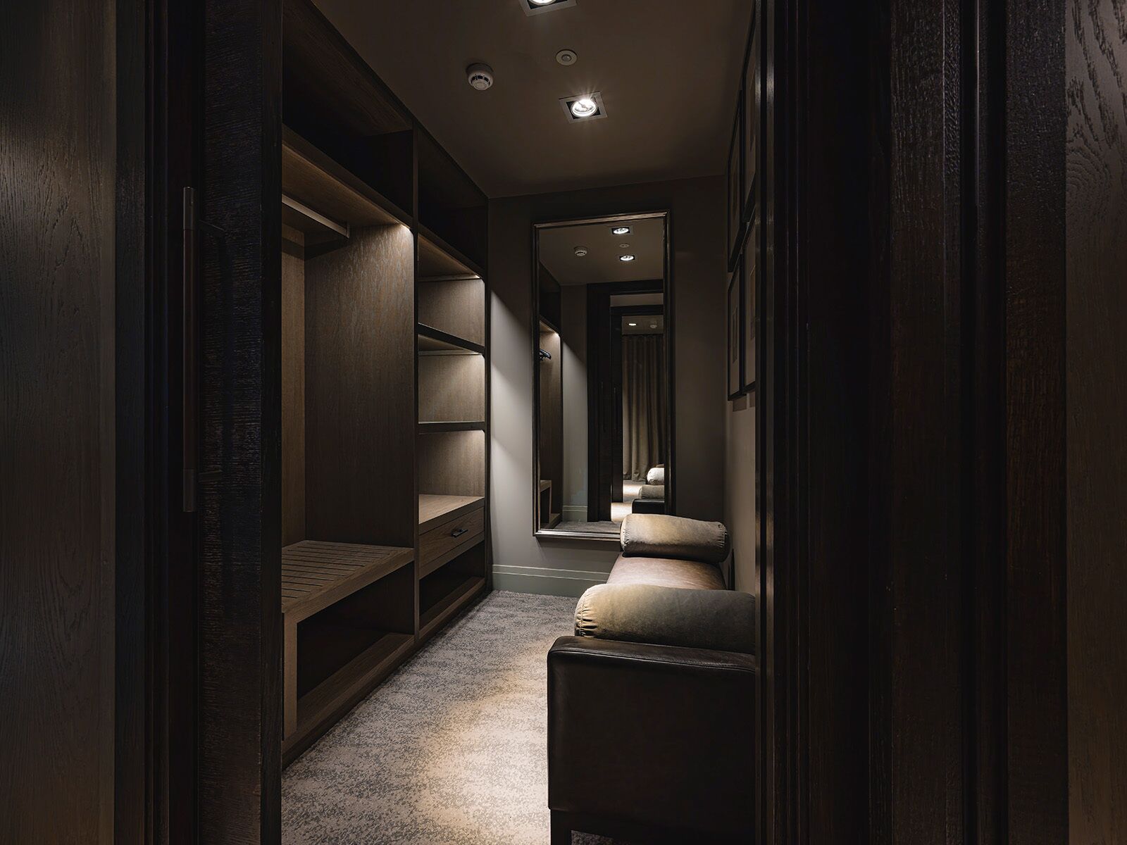 A narrow hallway with built-in shelving, a bench, and a mirror reflecting a bed.