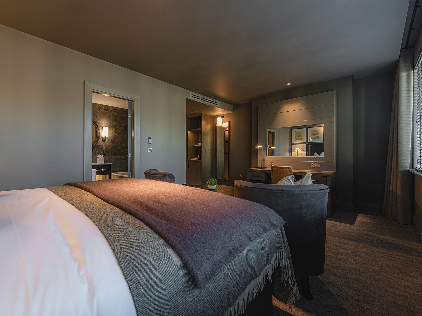 A modern hotel room with a large bed, a desk, a vanity with a mirror, and a bathroom visible through an open door.