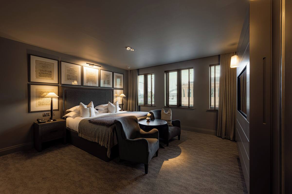 Una habitación de hotel moderna con una cama grande, dos sillones, una mesita y documentos enmarcados en la pared.