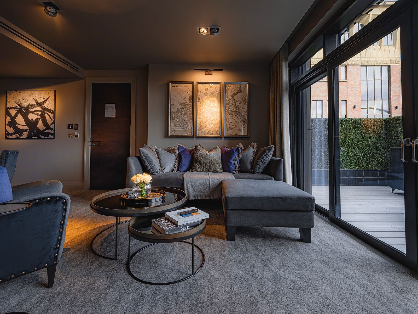 A modern living room with a grey sofa, a round coffee table with books, and a large window offering a view of a brick building and greenery.