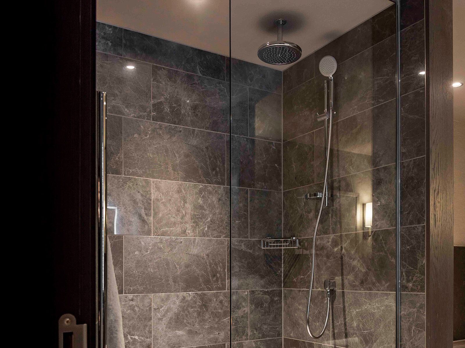 Une douche moderne, dotée d’un pommeau de douche à main et d’un robinet mural, le tout installé sur un mur recouvert de carreaux sombres évoquant le marbre.