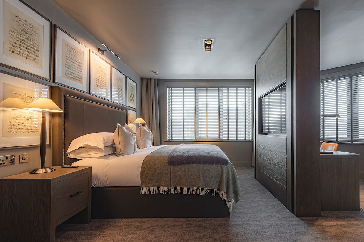 Habitación de hotel moderna con una cama grande, una mesita de noche, un escritorio y una ventana con persianas.