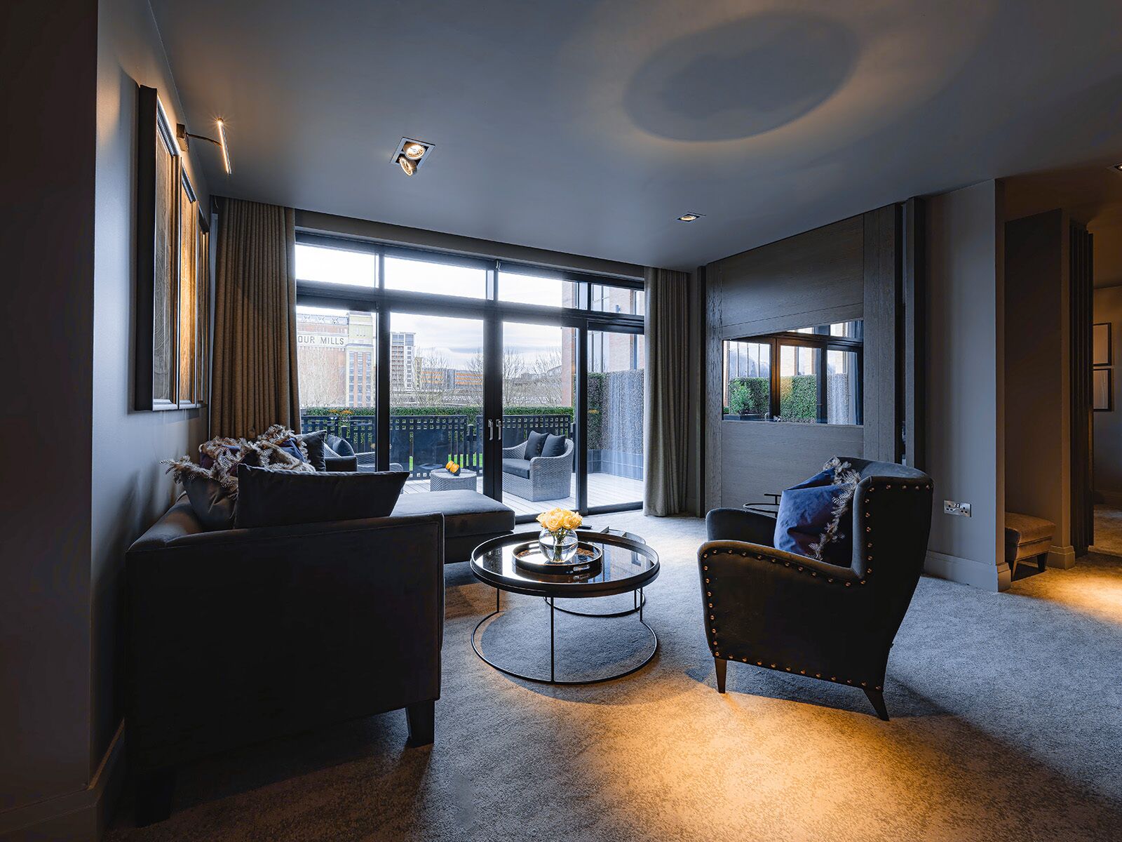 A modern living room with a sofa, armchair, and coffee table, large windows with a view of buildings, and a ceiling with recessed lighting.
