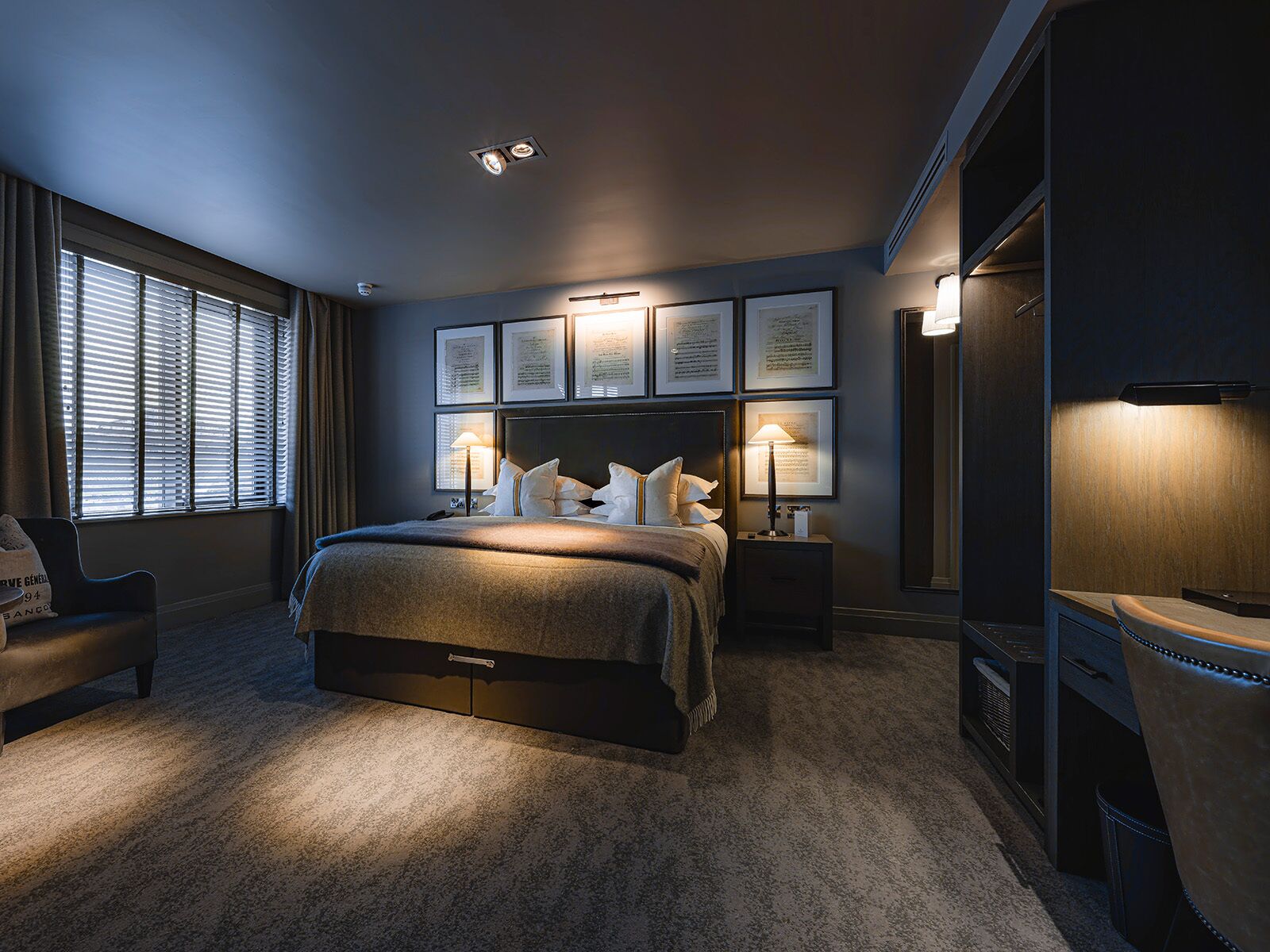 A modern hotel bedroom with a large bed, two bedside tables with lamps, a desk with a chair, and framed certificates on the wall.