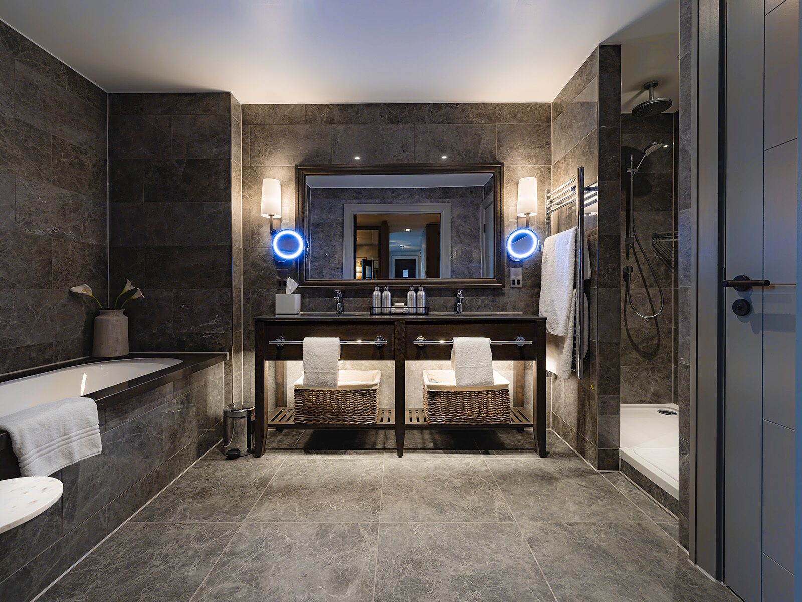 Un baño moderno con pared de mármol oscuro, un espejo grande, dos lavamanos con toalleros, una bañera y una ducha a ras de suelo.
