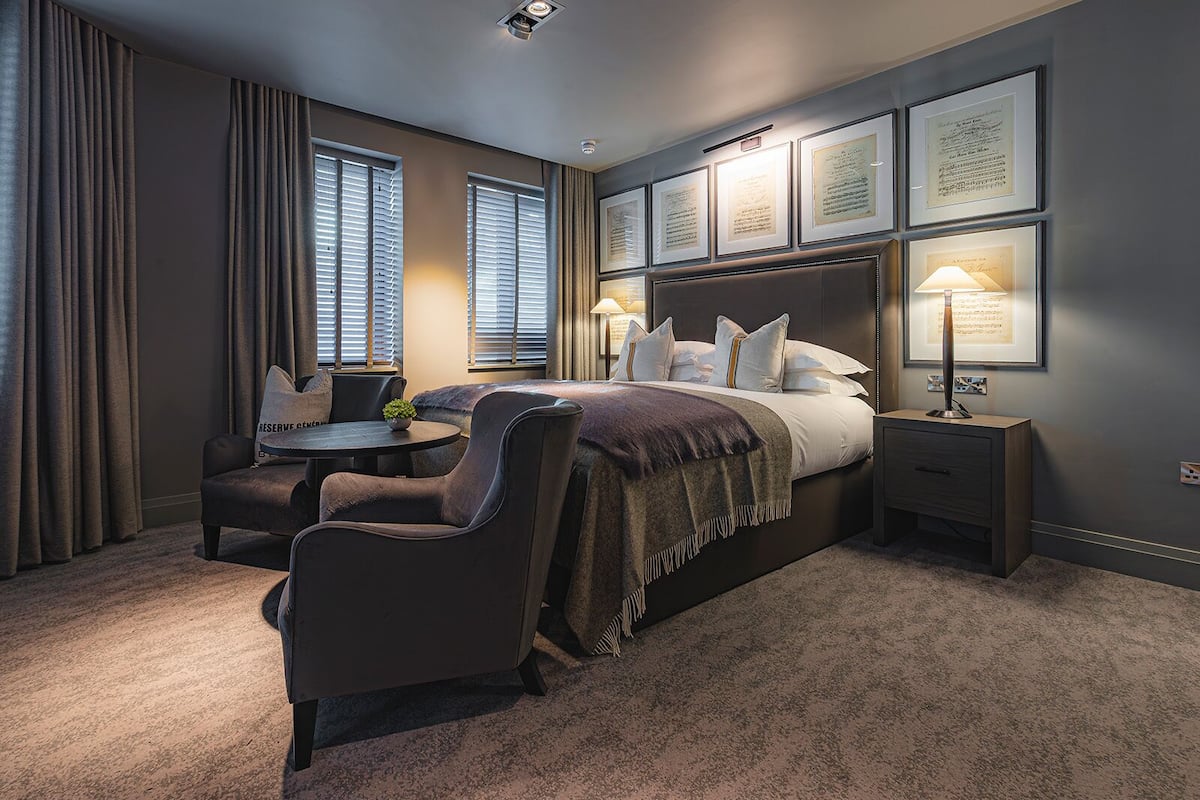 Una habitación de hotel moderna con una cama grande, una mesa redonda y una silla. Hay diplomas enmarcados en la pared y una lámpara sobre la mesita de noche.