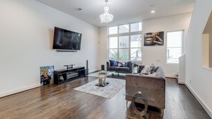 A spacious living room with hardwood flooring, a large TV mounted on the wall, a modern chandelier, and a large window providing natural light. - Posh Italian home with a rooftop terrace in the heart of Houston! (Houston)