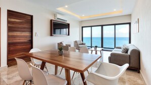 Un salón moderno con una mesa de comedor de madera, sillas blancas, un sofá y un amplio ventanal con vistas al mar.