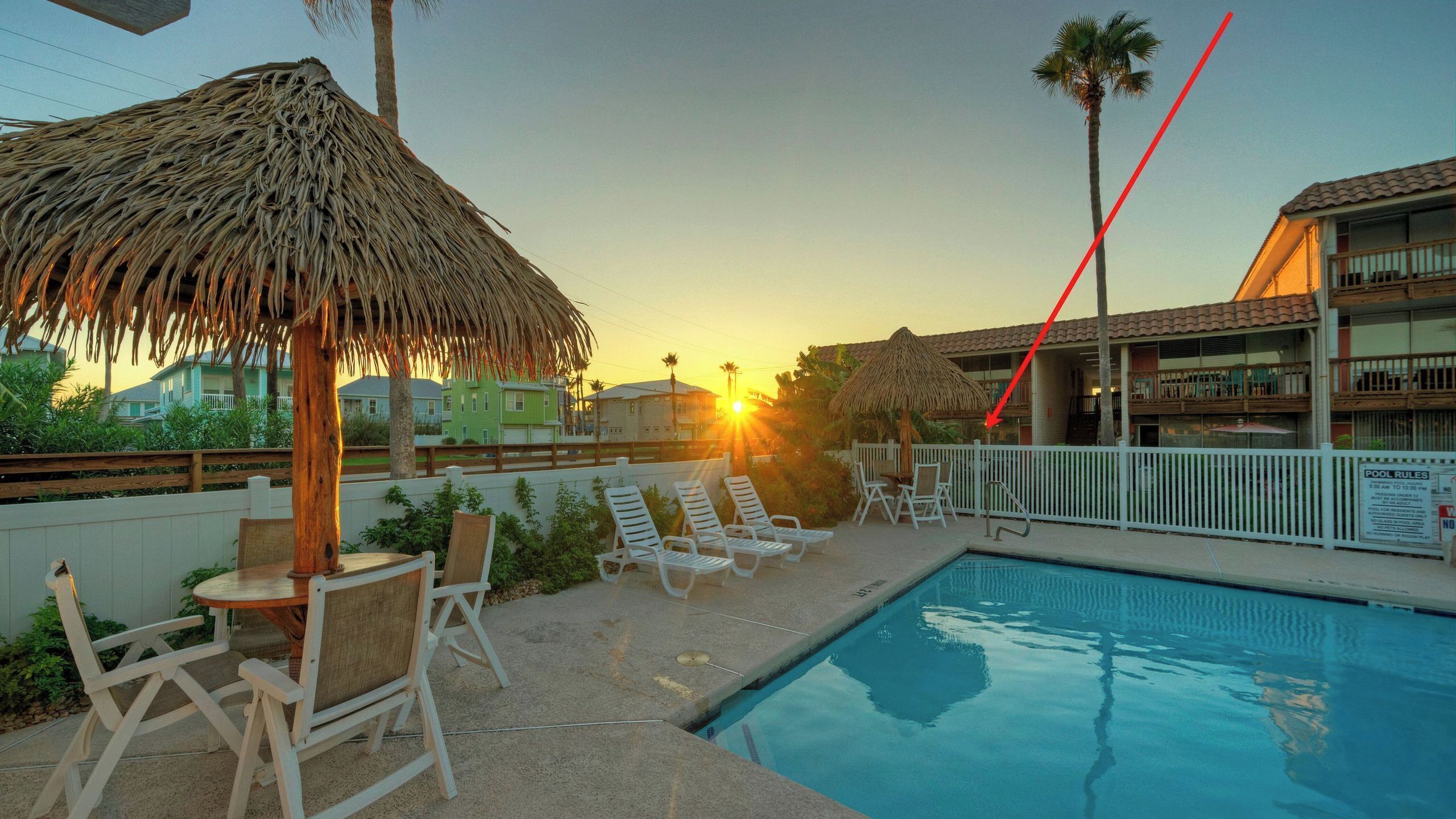 A pool area with lounge chairs and a thatched umbrella, surrounded by a fence and buildings in the background.
