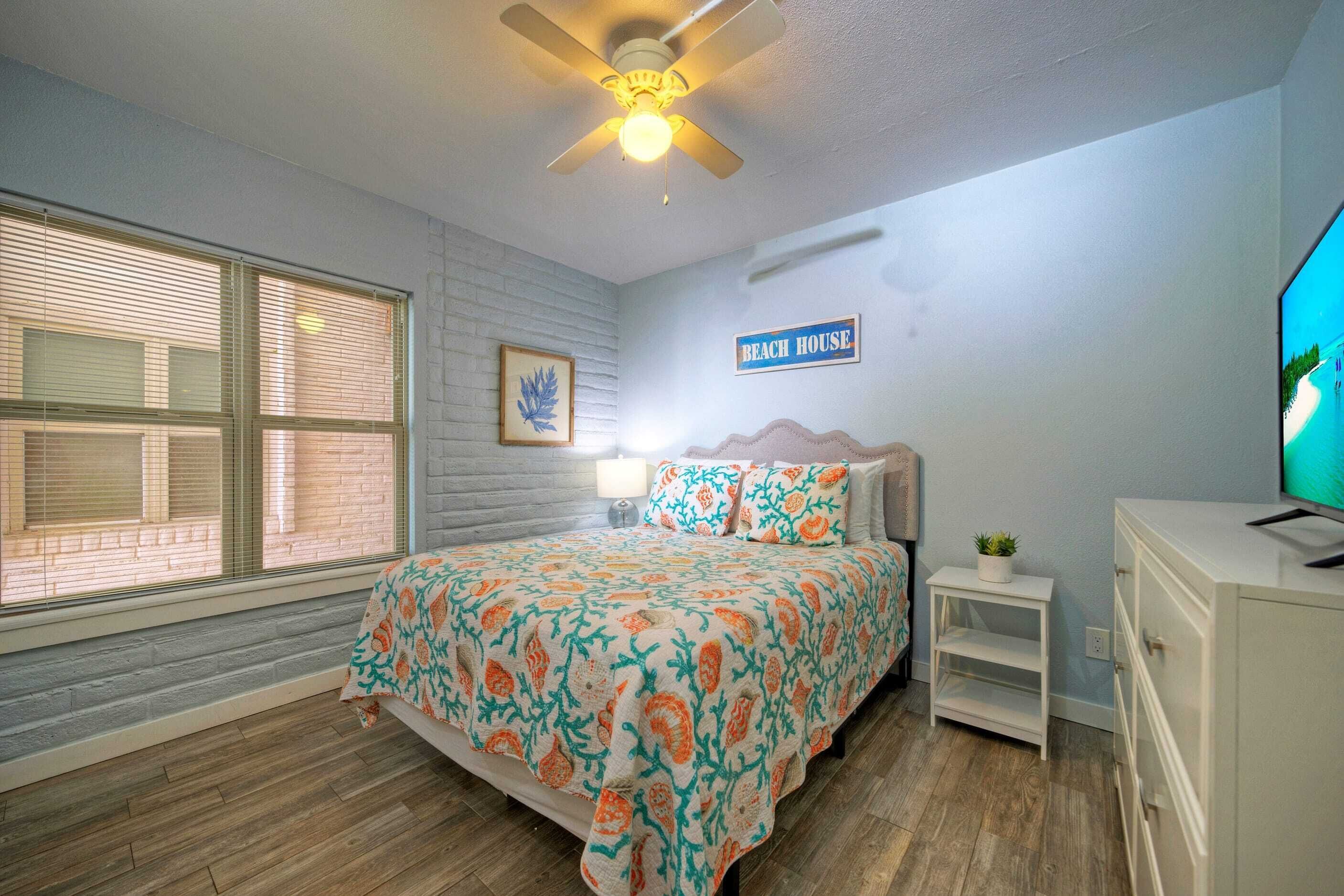 A bedroom with a bed, a ceiling fan, a TV, a nightstand, and a sign that reads "BEACH HOUSE".