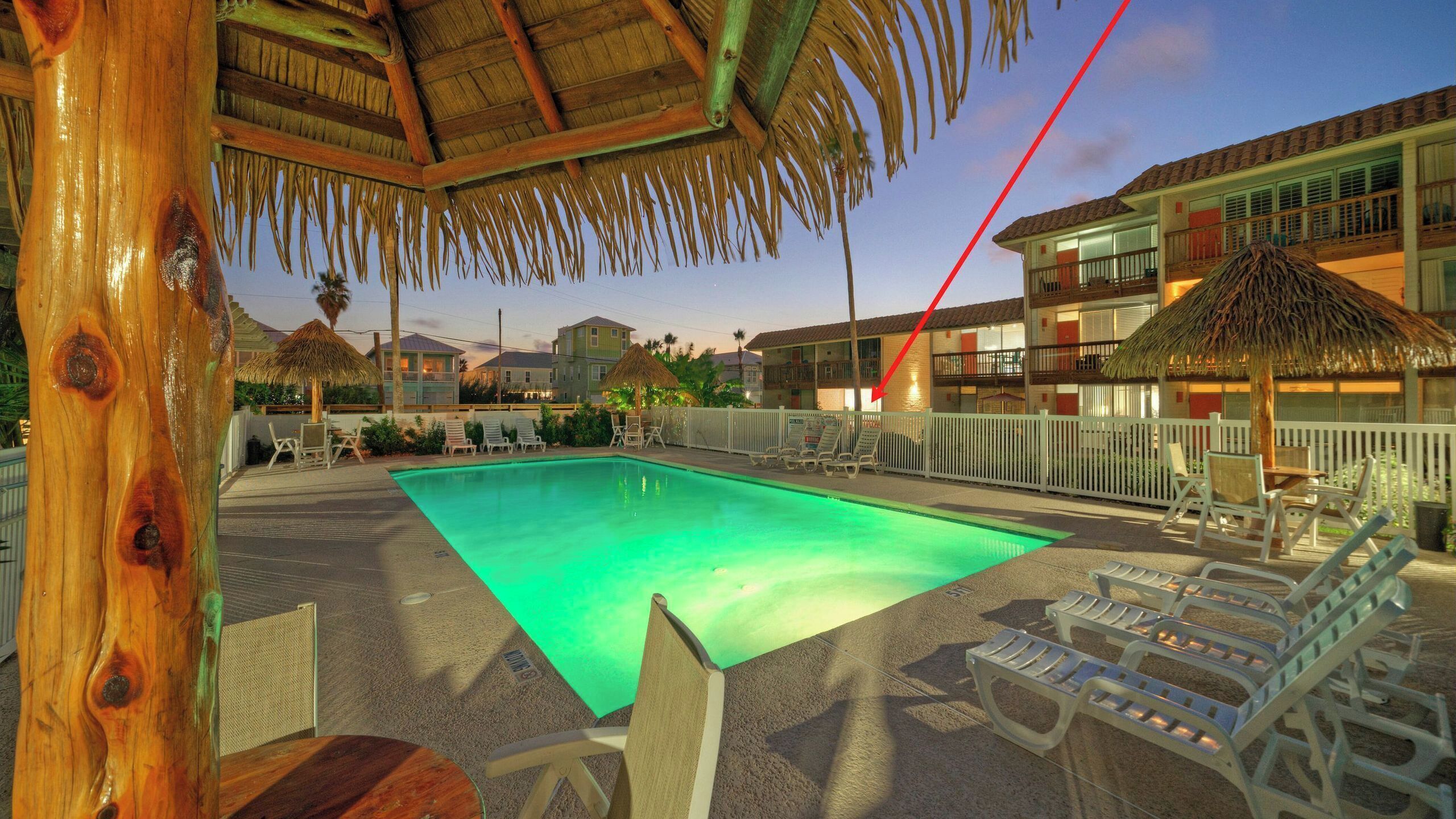 A swimming pool with lounge chairs, a thatched roof structure, and a multi-story building with balconies.
