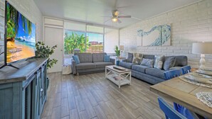 A living room with a sofa, coffee table, and TV. There is a window with blinds, a ceiling fan, and a brick wall. - Beach Boardwalk Access Pool + Remodeled Interior Tropical Haven by Avantstay (Port Aransas)