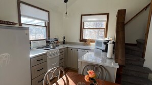 Eine kleine Küche mit weißen Geräten, einem Holztisch und Blick ins Freie durch ein Fenster.