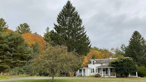 Ein weißes Haus mit Veranda, umgeben von einer grünen Wiese und Bäumen mit Herbstlaub.