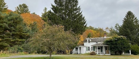 A white house with a porch, surrounded by a grassy lawn and trees with autumn foliage.