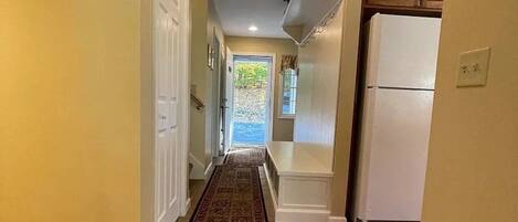 A hallway with a tiled floor, a door on the left, a window with curtains, and a refrigerator on the right.