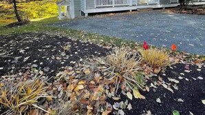 Una casa de dos pisos con un porche delantero, un pequeño jardín con hojas otoñales y un cielo azul despejado.